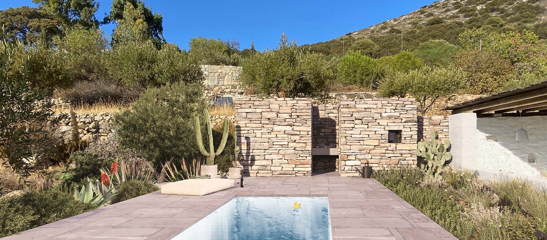 Vista frontal de piscina en villa griega, con un volumen de piedra al fondo integrado con el muro de contencion del terreno.